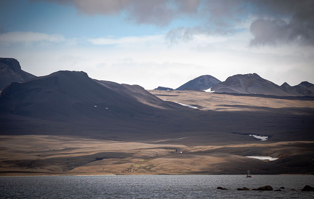 Rugged Icelandic landscape with mountains, snow patches, and a body of water under an overcast sky.