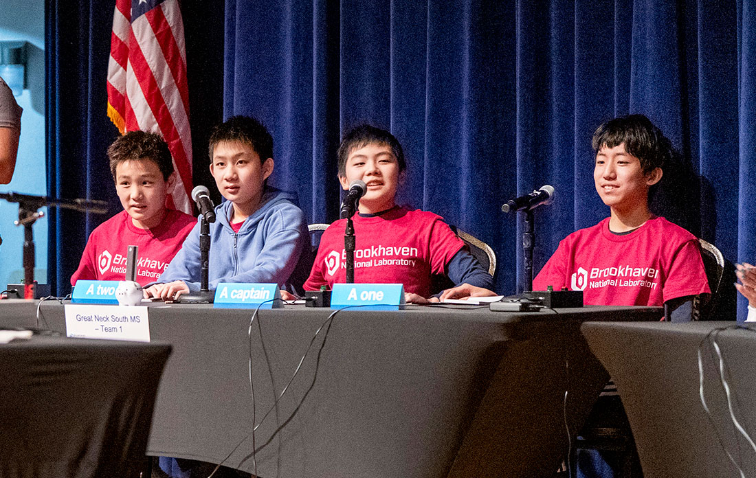 Students wearing Brookhaven National Laboratory T-shirts sit behind microphones on a stage during a